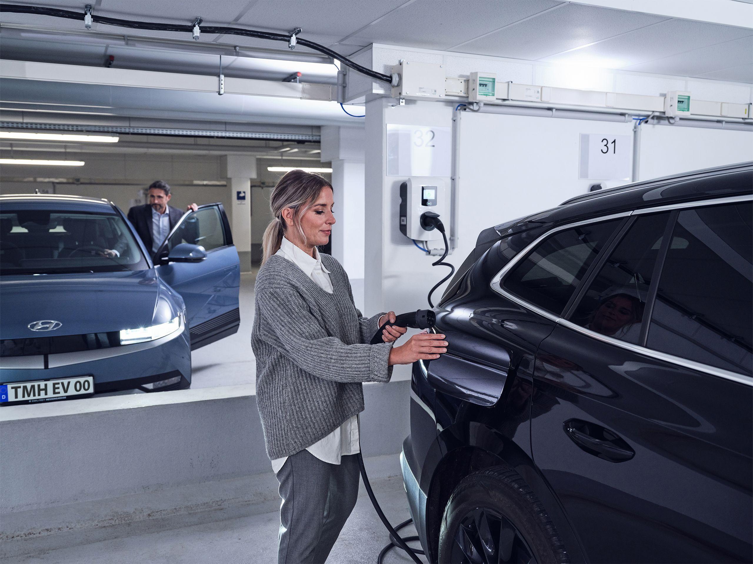 Frau steckt Ladekabel am Elektroauto an mit Kollege im Hintergrund